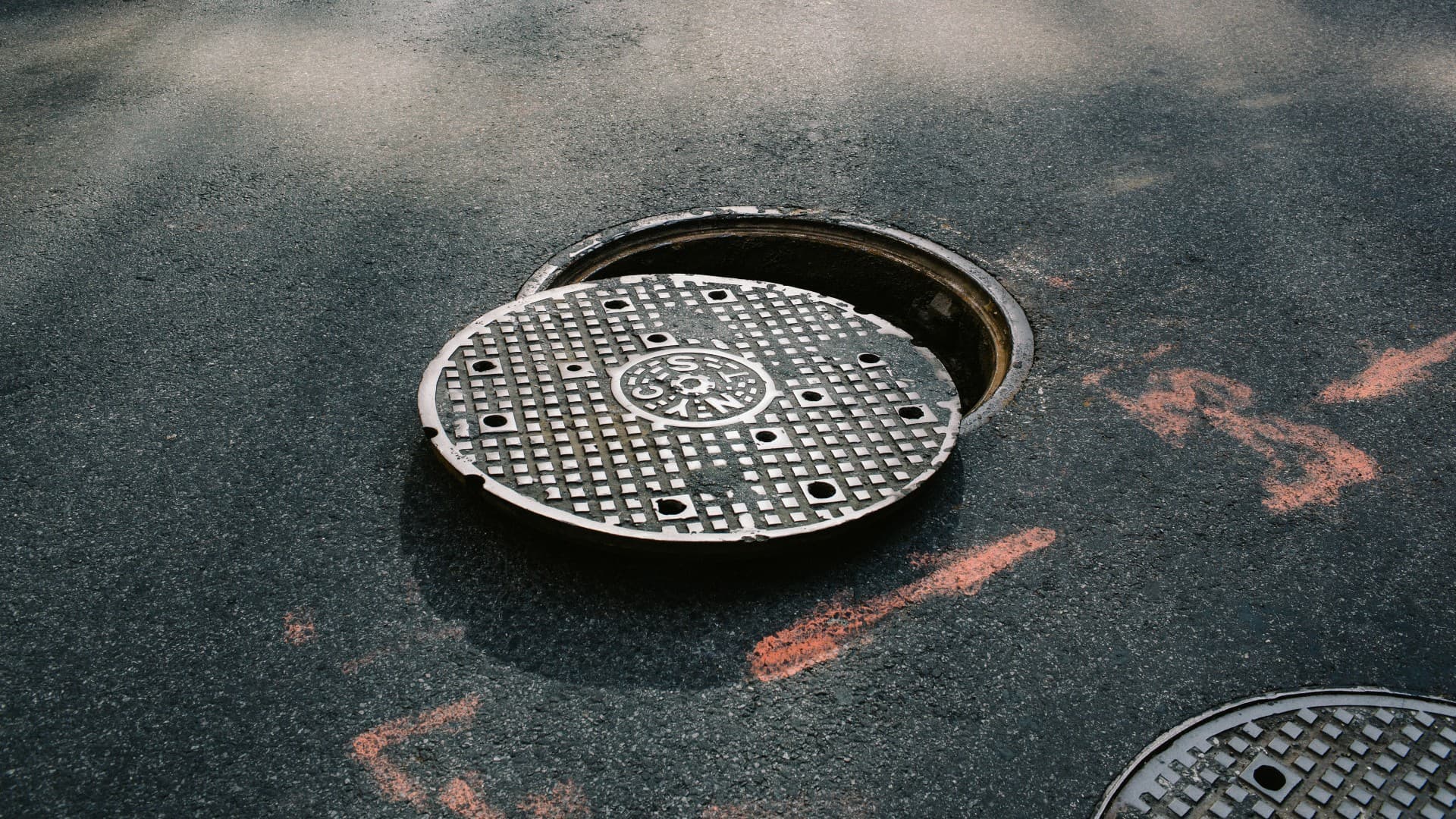 A cast iron manhole cover set into city pavement, showing its circular shape and textured grip surface
